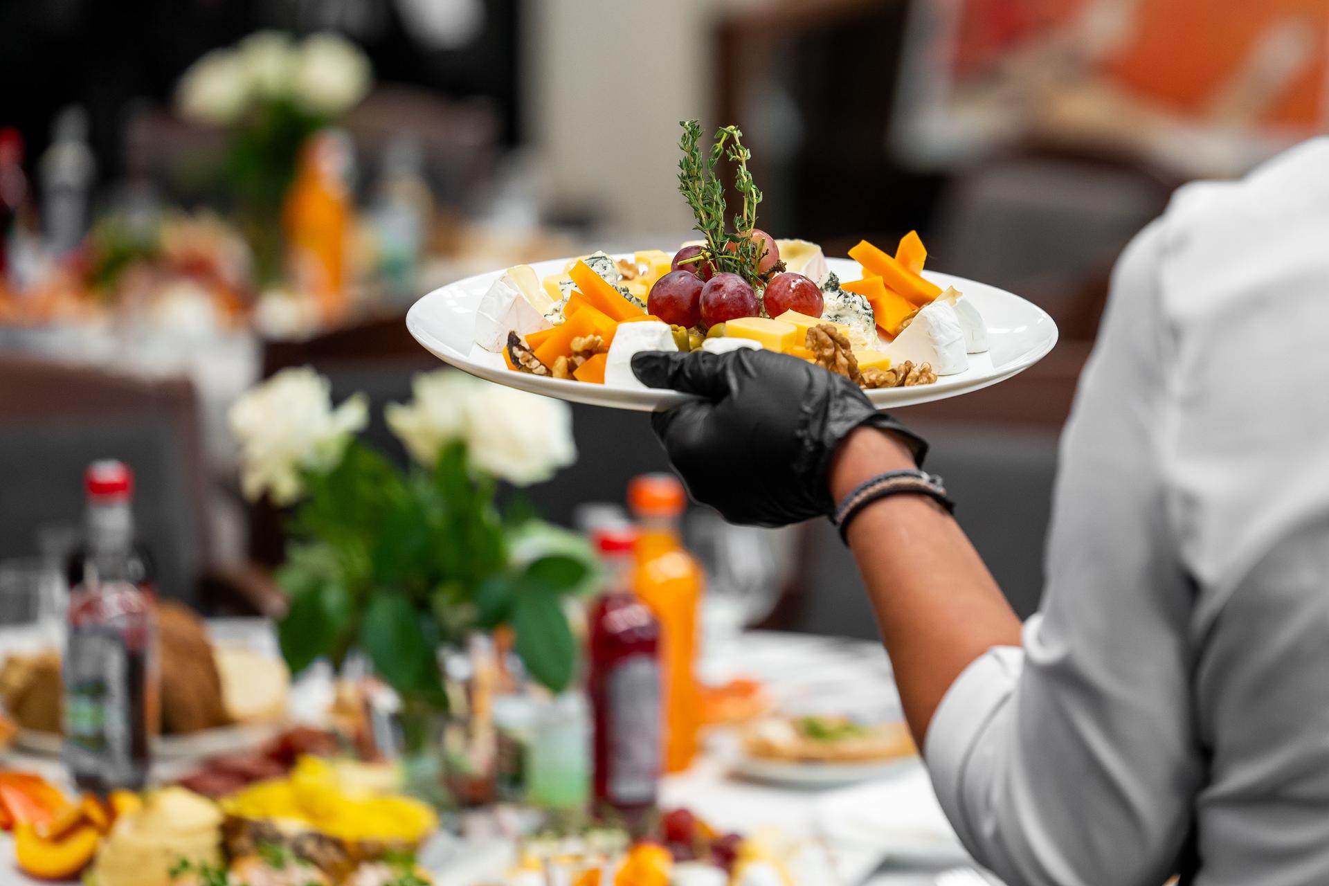 Waiter in black gloves serving gourmet cheese plate with grapes, nuts, and herbs at elegant event table with drinks and floral decor. Close-up, catering concept.